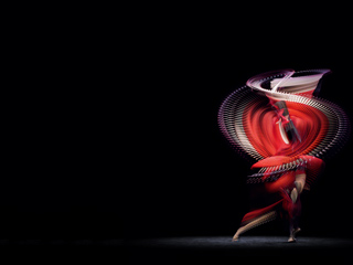 A dancer spins through a dark space, wearing a brilliant red dress which leaves traces through the air as she moves.