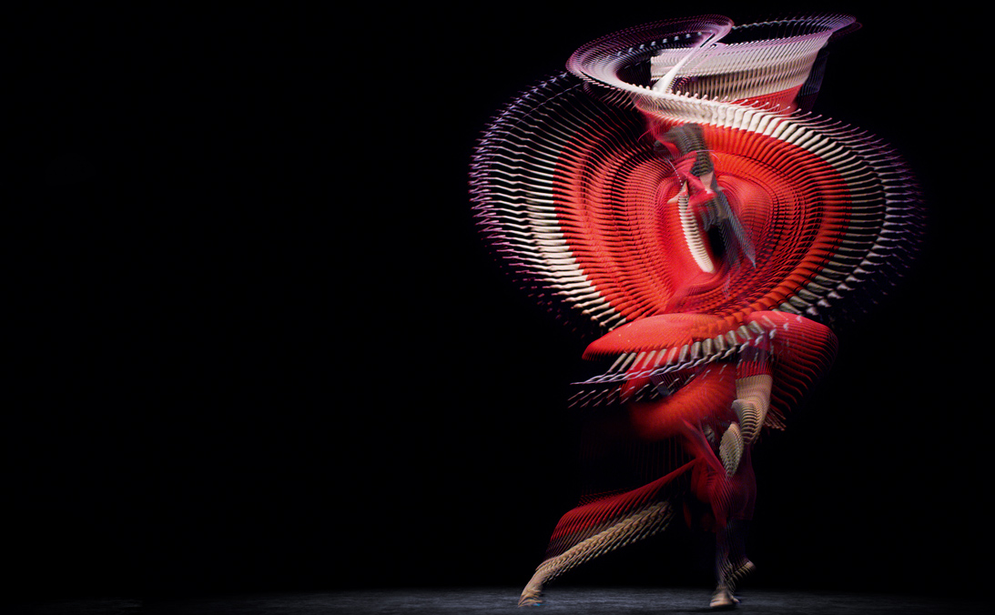A dancer spins through a dark space, wearing a brilliant red dress which leaves traces through the air as she moves.